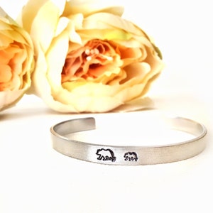 Könnte beinhalten: Ein silbernes Armband mit zwei eingravierten Bärensilhouetten. Das Armband liegt auf einer weißen Oberfläche mit einer gelben Blume im Hintergrund.