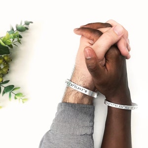 May include: Two people holding hands, each wearing a silver bracelet engraved with coordinates. The bracelets are a simple and elegant way to commemorate a special place or date.