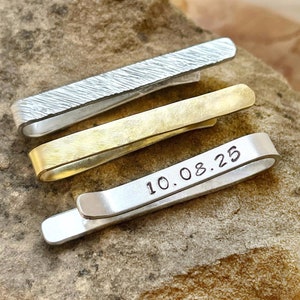 May include: Three metal tie clips in silver, gold, and silver. The top clip has a textured pattern, the middle is gold, and the bottom silver clip has the date "10.08.25" engraved on it. The clips are displayed on a rough, brown stone surface.
