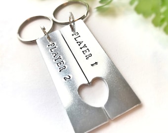 Op de afbeelding: Twee zilveren sleutelhangers met een hart uitgesneden in het midden. De sleutelhangers zijn gegraveerd met de woorden "Player 1" en "Player 2".