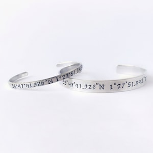 May include: Two silver bracelets with engraved coordinates. The top bracelet reads "56° 47'41.926"N 1°27'51.843" and the bottom bracelet reads "53° 47'41.926"N 1°27'51.843"T".