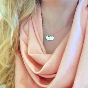 May include: A silver necklace with three round charms, each with a letter engraved on it. The necklace is worn by a person with blonde hair and a peach-colored scarf.