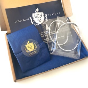 May include: A blue and white gift box with a gold logo that says "Goldcrest Designs" and a gold crest with the letters "GD" inside. The box is open and contains a blue tissue paper with a sticker that says "Goldcrest Designs" and a clear plastic bag with two silver bracelets.