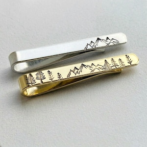 May include: Two metal tie clips, one silver and one gold. Both feature a mountain and tree design. The silver clip has a mountain range etched on it, while the gold clip has a forest and mountain range design.