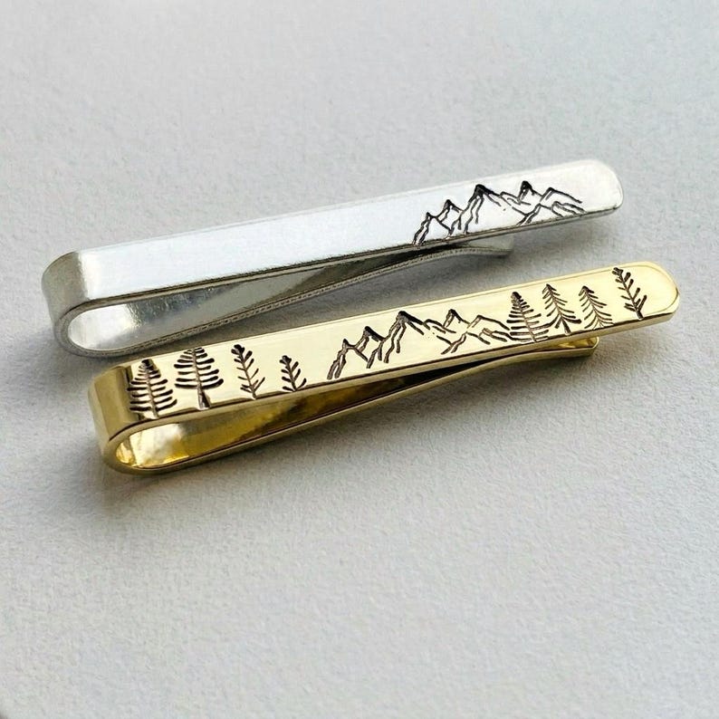 May include: Two metal tie clips, one silver and one gold. Both feature a mountain and tree design. The silver clip has a mountain range etched on it, while the gold clip has a forest and mountain range design.