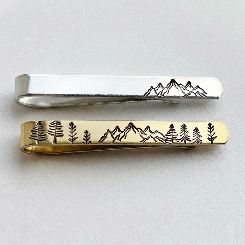 May include: Two metal tie clips. One is silver-toned with a mountain range design in black. The other is gold-toned with a forest and mountain range design in black. Both clips have a curved, spring-loaded design.
