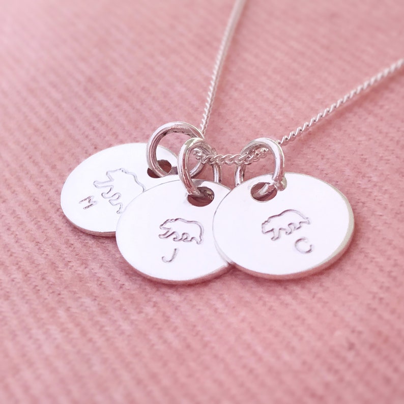 May include: Silver necklace with three round charms featuring a bear silhouette and the letters M, J, and C.