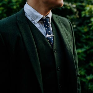 May include: A dark green suit with a matching vest and a light blue collared shirt. A navy blue tie with a pattern of small, light-colored shapes is secured with a silver tie clip. The suit is buttoned, and the background is a blurred green.