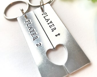 Op de afbeelding: Twee zilveren sleutelhangers met de woorden "Player 1" en "Player 2" erop gegraveerd. De sleutelhangers zijn verbonden door een hartvormige uitsparing.