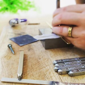 May include: A close-up of a person using a metal hammer to stamp letters into a metal surface. The surface is covered in a white paper. There are several metal stamps on the table, each with the word "ImpressArt" engraved on them.