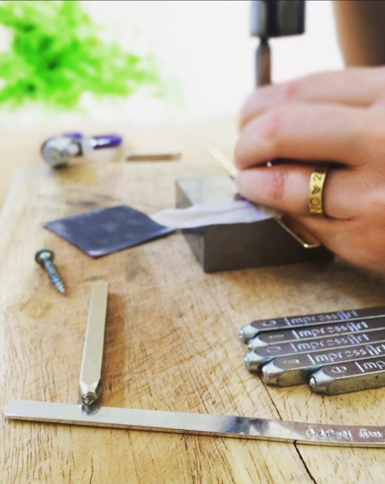 May include: A close-up of a person using a metal hammer to stamp letters into a metal surface. The surface is covered in a white paper. There are several metal stamps on the table, each with the word "ImpressArt" engraved on them.