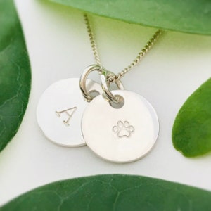 May include: Silver necklace with two charms. One charm is a round disc with the letter 'A' engraved on it. The other charm is a round disc with a paw print engraved on it.
