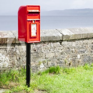 May include: A bright red mailbox stands on a black post, set against a stone wall and green grass. The mailbox has a slot for mail and a small window. The background shows a body of water.