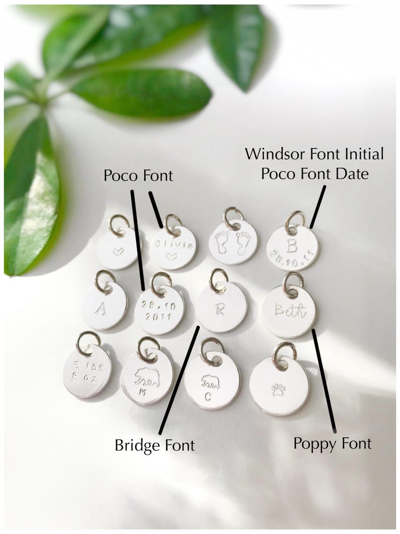 May include: A collection of silver charms with different fonts and designs. The charms include a heart, a footprint, a bear, a paw print, and the letters A, B, R, and the names Olivia and Beth. The date 28.10.11 is also included on one of the charms. The charms are labeled with the fonts Poco, Windsor, Bridge, and Poppy.