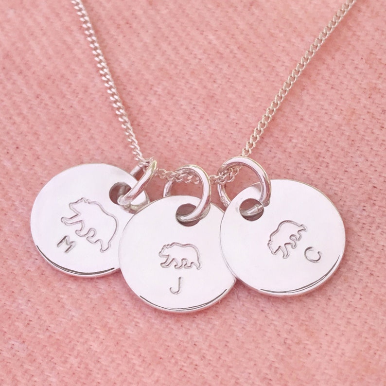 May include: Silver necklace with three round charms featuring bear silhouettes and the letters M, J, and C.
