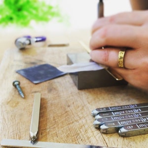 May include: A close-up of a metal stamping tool set with the word "ImpressArt" engraved on each tool. The tools are arranged on a wooden surface. A silver metal ruler is also visible in the image.