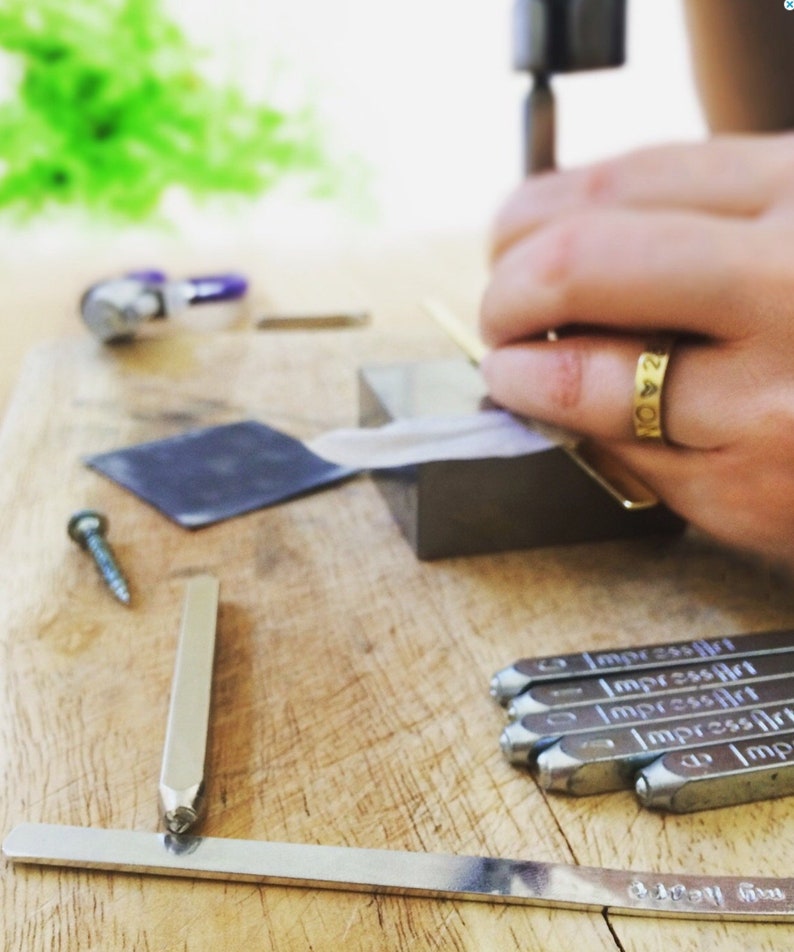 May include: A close-up of a metal stamping tool set with the word "ImpressArt" engraved on each tool. The tools are arranged on a wooden surface. A silver metal ruler is also visible in the image.