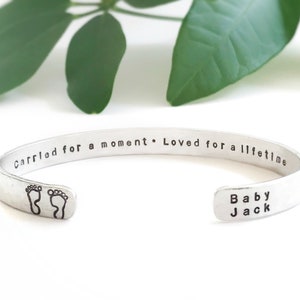 May include: Silver cuff bracelet with the inscription "Carried for a moment. Loved for a lifetime." and the words "Baby Jack" on the other side. The bracelet also features a pair of footprints.