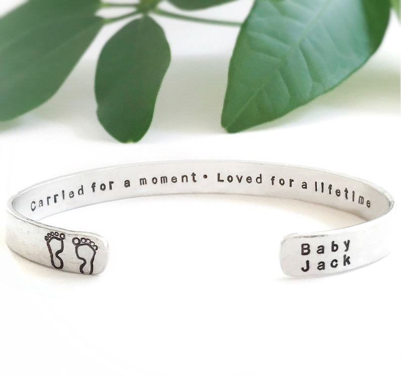 May include: Silver cuff bracelet with the inscription "Carried for a moment. Loved for a lifetime." and the words "Baby Jack" on the other side. The bracelet also features a pair of footprints.