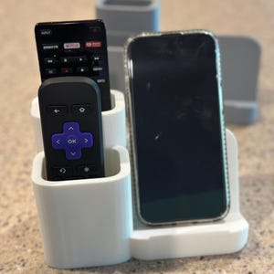 May include: A white plastic organizer holds two black remote controls and a smartphone. The remotes have the Amazon, Netflix, and Roku logos. The smartphone has a clear case.