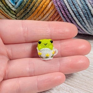May include: A tiny, handmade crochet frog in a teacup. The frog is lime green with black eyes, and the teacup is white with yellow flower accents. The frog is sitting in a white teacup with yellow flowers. The background shows a skein of yarn.