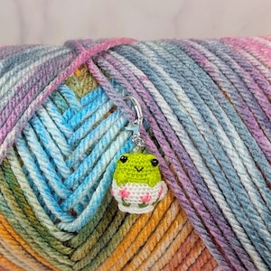 May include: A small, crocheted frog stitch marker featuring a bright green head with black bead eyes and a white base decorated with tiny pink flowers. The frog is attached to a silver-toned metal lobster clasp hook for easy attachment to yarn.