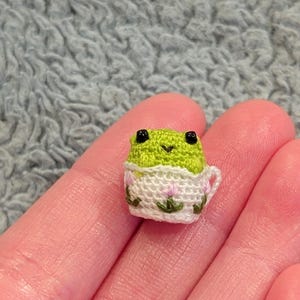 May include: A tiny, handmade crochet frog in a teacup. The frog is green with black eyes, and the teacup is white with pink floral accents. The teacup is held in a person's hand.