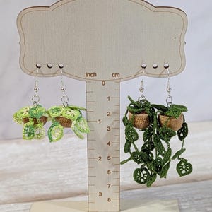 May include: Two pairs of dangling earrings featuring miniature crocheted potted plants. The left pair depicts light green variegated foliage in tan pots, while the right pair features dark green trailing leaves in tan pots, all on silver-toned hooks.