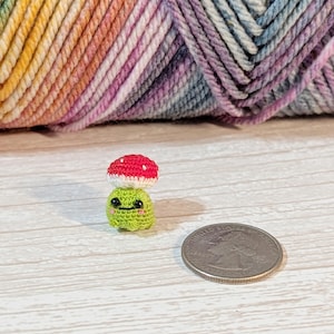 May include: A miniature crocheted mushroom figurine with a bright red cap featuring white spots and a light green base with a small embroidered face. The tiny object is positioned next to a standard United States quarter coin for scale.