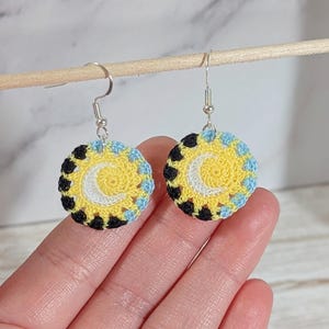 Peut inclure: Une paire de boucles d'oreilles crochetées à la main. Chaque boucle d'oreille présente un centre jaune avec un croissant de lune blanc, entouré de crochet noir, bleu et jaune. Les boucles d'oreilles ont des crochets d'oreille argentés.