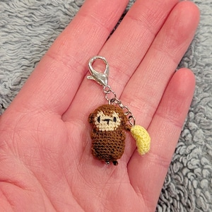 May include: A small, crocheted monkey charm with a yellow banana. The monkey is brown with a cream-colored face and a silver clasp. The charm is handmade and appears to be a keychain or bag accessory.