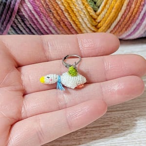 May include: A small, white, crocheted goose stitch marker featuring a yellow beak, a light blue scarf, and a tiny green accessory on its back. The goose is attached to a silver-toned metal lever-back clasp, designed for use in knitting or crochet projects.