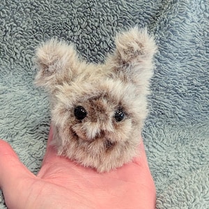 May include: A small, plush, light brown creature with black eyes and a smiling expression. The furry toy has two small ears and is held in a hand against a textured, light blue background.