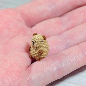 May include: A miniature crochet capybara figurine. The small, handmade animal is primarily tan with darker brown accents on its ears, feet, and face. The capybara has closed eyes, suggesting it is sleeping. The figurine is small enough to fit in the palm of a hand.