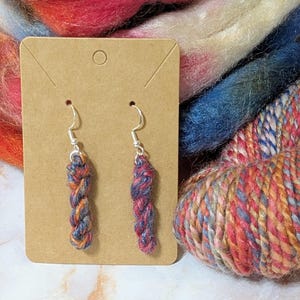 May include: A pair of drop earrings featuring small, braided segments of multi-colored yarn in shades of blue, orange, and red. The yarn segments are attached to silver-toned metal fishhook ear wires and are displayed on a rectangular brown card.