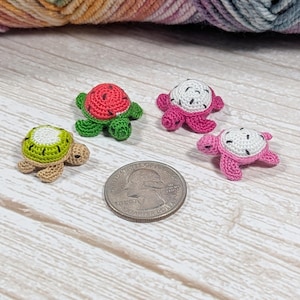 May include: Four miniature crocheted turtles with fruit-themed shells, including watermelon, kiwi, and dragon fruit designs, arranged around a US quarter coin for scale, showcasing intricate yarn work and vibrant colors on a light wood surface.
