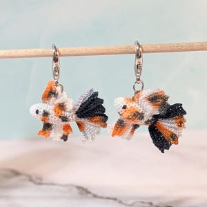 May include: A pair of dangling earrings featuring miniature crocheted betta fish charms. Each fish is crafted with intricate thread work in a calico pattern of white, orange, and black, suspended from silver-toned metal lobster claw clasps.