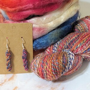 Peut inclure: Une paire de boucles d'oreilles pendantes en forme de mini &eacute;cheveaux de laine aux nuances vari&eacute;es de rouge, bleu et orange, fix&eacute;es sur des crochets argent&eacute;s sur un support en carton brun, pr&eacute;sent&eacute;es avec un &eacute;cheveau de laine et de la laine card&eacute;e.