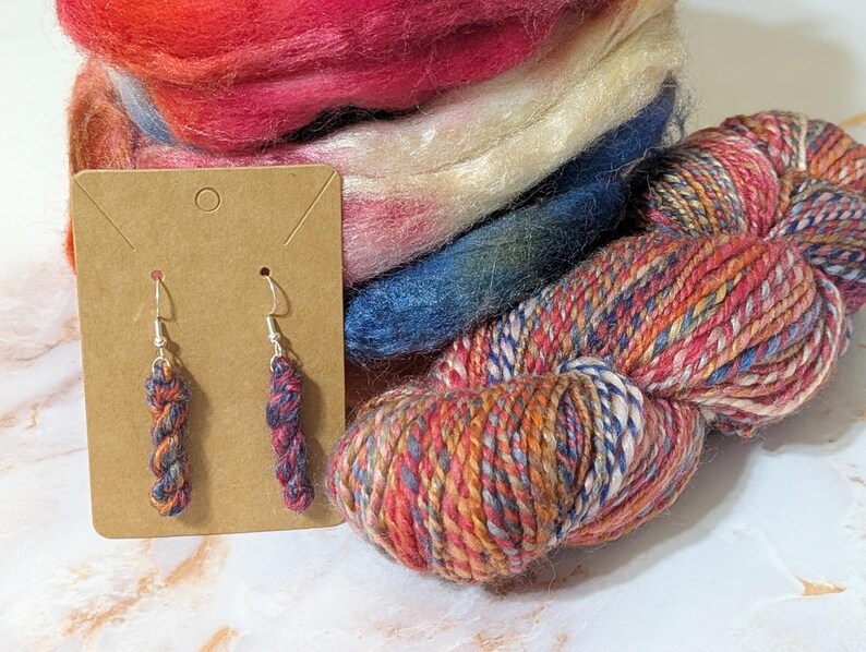 Peut inclure: Une paire de boucles d'oreilles pendantes en forme de mini &eacute;cheveaux de laine aux nuances vari&eacute;es de rouge, bleu et orange, fix&eacute;es sur des crochets argent&eacute;s sur un support en carton brun, pr&eacute;sent&eacute;es avec un &eacute;cheveau de laine et de la laine card&eacute;e.