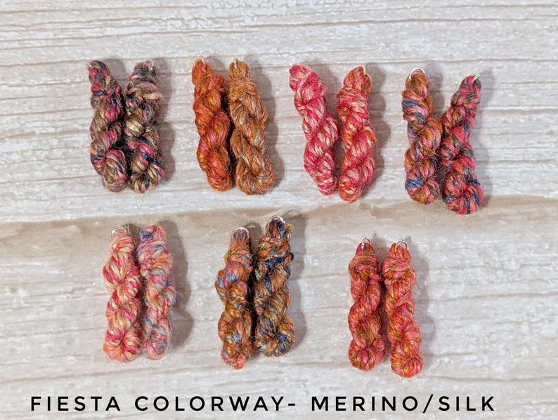 Peut inclure: Sept paires de petits &eacute;cheveaux de fil torsad&eacute;s dans divers coloris panach&eacute;s, incluant des nuances de rose, orange, marron et bleu. Le texte en bas indique FIESTA COLORWAY- MERINO/SILK. Chaque &eacute;cheveau pr&eacute;sente un aspect textur&eacute; et multicolore.