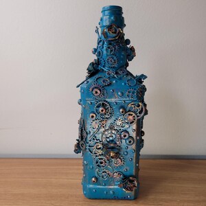 May include: A blue glass bottle decorated with gears, cogs, and other steampunk-inspired elements. The bottle has a gold metallic finish and is accented with small, decorative details.