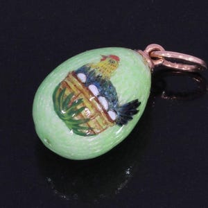 Può includere: Un ciondolo a forma di uovo verde chiaro con un anello dorato. Il ciondolo presenta una scena dipinta di un gallo in un cesto con uova. Il gallo ha dettagli rossi, gialli, blu e neri. Il cesto è marrone e verde.