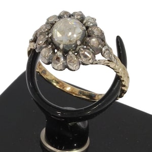Puede incluir: Un anillo vintage con una gema grande, redonda y transparente, rodeada de piedras más pequeñas en un diseño floral. El anillo tiene una banda dorada y un engaste plateado. El anillo se exhibe sobre un soporte negro.