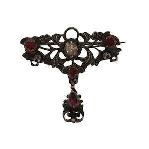 May include: A silver brooch with a delicate floral design and red and clear gemstones. The brooch has a dangling drop with a red gemstone.