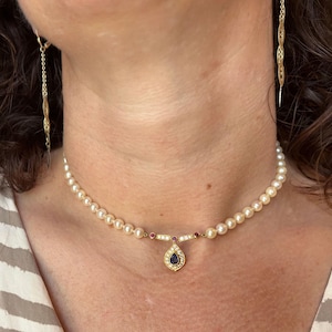 May include: A pearl necklace with a gold pendant featuring a teardrop-shaped sapphire and small diamonds. The necklace is accented with red gemstones and is paired with matching gold earrings.