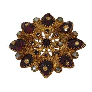 Puede incluir: Broche dorada con diseño de estrella. Presenta grandes piedras preciosas de color rojo oscuro y pequeños detalles claros y dorados. El intrincado trabajo de filigrana metálica añade detalles a esta joya de estilo vintage.