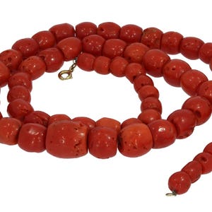 Puede incluir: Un collar compuesto por vibrantes cuentas de coral rojo. Las cuentas varían en tamaño y forma, con una mezcla de formas redondeadas y más rectangulares. Un cierre dorado asegura el collar, que se muestra sobre un fondo blanco.