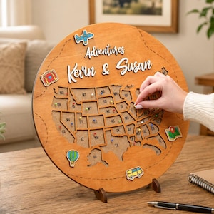 May include: A circular wooden map of the United States with a cork backing, featuring the text "Adventures" and "Kevin & Susan". The map includes small colorful push pins in various states and decorative wooden icons of a plane, suitcase, and van.