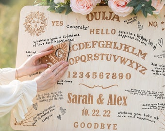 May include: A wooden Ouija board with a planchette on top. The board has a sun and moon design and the letters of the alphabet, numbers 1-9 and 0, and the words "YES", "NO", "GOODBYE", "HELLO", "CONGRATS!" and "OUIJA". The board is signed "Sarah & Alex" and "10. 22. 22".