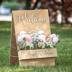 Puede incluir: Un letrero de madera de bienvenida con un arreglo floral en una maceta. El letrero dice "Bienvenido" y "Ainsley & Sherine" en cursiva.